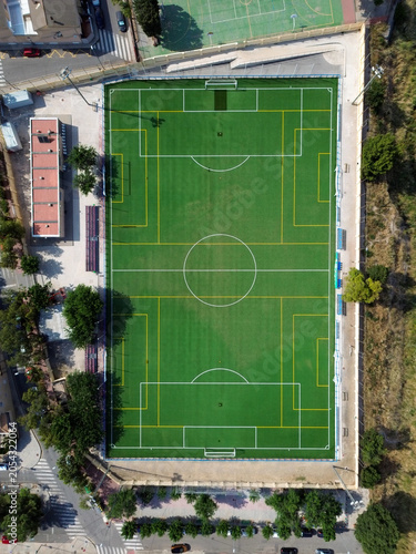 Vertical drone perspective of a vibrant green multi sport stadium field surrounded by stands buildings streets and trees in a coastal city neighborhood under bright sunny daylight conditions