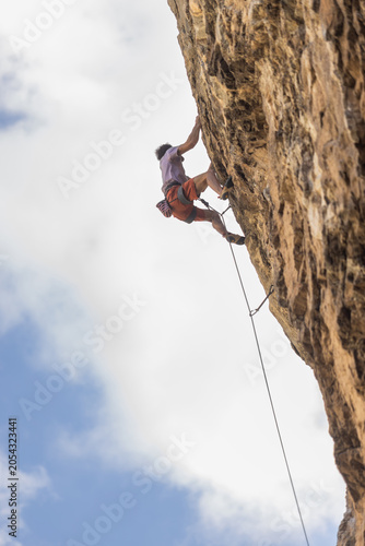 Climber ascends a steep rock face. They grip cracks with focused concentration. White helmet and harness secure their position. Blue sky and soft clouds frame the scene