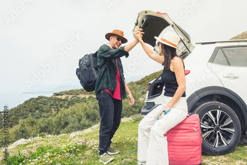 Fotografie Happy Travelers tourist couple hi5 stand put luggage to white rented SUV vehicle trunk on scenic Mediterranean viewpoint