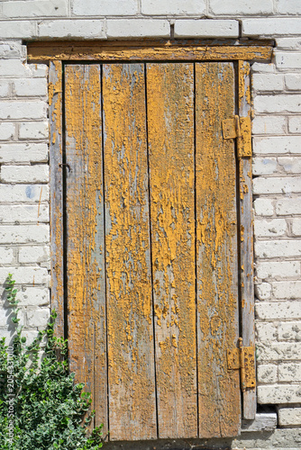 Ancient weathered wooden door with peeling yellow paint in a white brick wall.