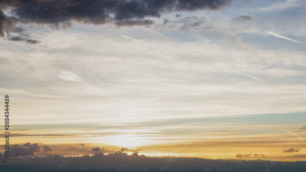Fototapeta premium Panoramic view of a dramatic sunset sky with layered clouds illuminated by warm golden light. Atmospheric evening landscape featuring colorful cloud formations, soft sunlight and a distant horizon. Pe