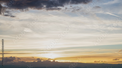 Panoramic view of a dramatic sunset sky with layered clouds illuminated by warm golden light. Atmospheric evening landscape featuring colorful cloud formations, soft sunlight and a distant horizon. Pe