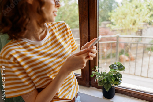 Photos A young brunette woman in a striped tee stands by a sunlit window, glancing at her phone with a small plant on the sill and a calm, casual mood nearby