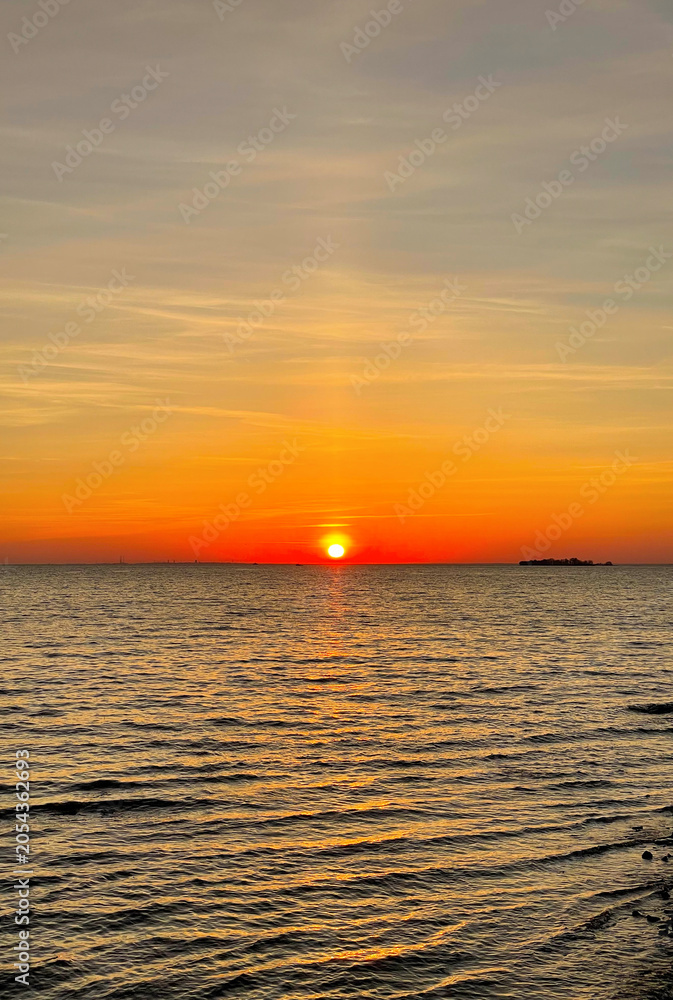 Obraz premium Sun dips below horizon over calm waters, casting warm orange glow across sky and sea. Serene sunset marks end of day.