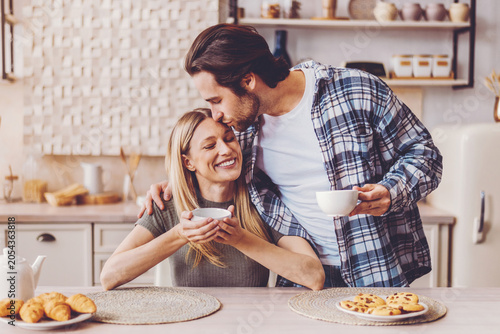 Quadro su tela A man kisses a woman on the forehead while she holds a bowl in a kitchen