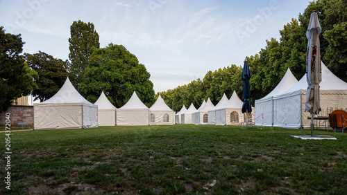 Row of white event tents set up on a green lawn surrounded by trees in a public park. Outdoor venue prepared for a festival, fair, exhibition, wedding or corporate event. Peaceful evening atmosphere w