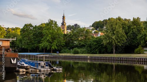 Scenic riverside view with small boats moored along the waterfront, lush green trees and a historic church tower rising above the town. Warm evening sunlight reflects on calm water, creating a peacefu
