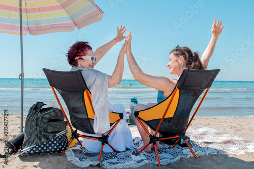 An elderly and young woman relax in sun loungers under an umbrella, holding hands by the sea. Safety, protection from the sun, heatstroke in the heat. Accessories for summer. Intergenerational