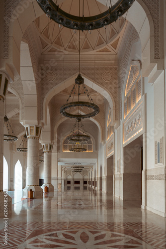 Architectural interior with ornate chandeliers, columns, arches, reflective marble floor, and decorative patterns.
