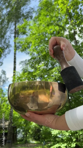 Spiritual wellness atmosphere with woman showing sound therapy instruments and singing bowls outdoors near green leaves and bright natural sky.