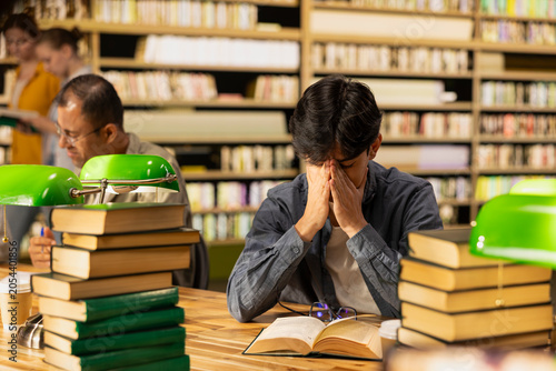 Εκτύπωση καμβά Overwhelmed male student rubbing eyes above open book in library, surrounded by stacks of books and green lamps during stressful exam preparation and fatigue, finals pressure, for study concepts