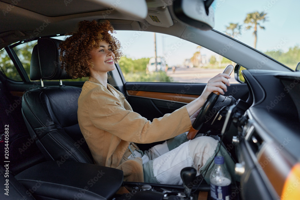 Naklejka premium Smiling woman with curly hair driving a car, wearing a beige jacket. Close up on interior with natural light. Concept of lifestyle, travel, and self expression. Retro film color adds warmth.
