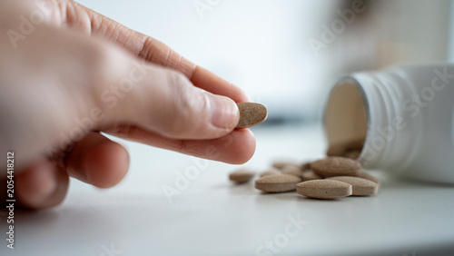 Hand picking single brown supplement tablet from white surface near open bottle