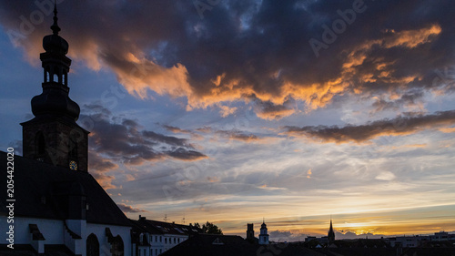 Historic church tower and old town skyline silhouetted against a dramatic sunset sky with glowing clouds and warm evening light. Colorful twilight atmosphere over a European city creates a scenic urba