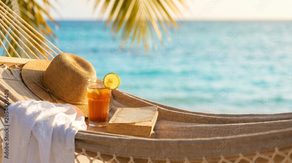 Obraz premium Relaxing tropical vacation scene with straw hat iced tea and book on hammock overlooking turquoise ocean for summer travel concept