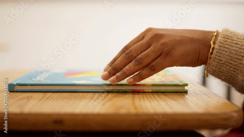 A woman's hand rests on a closed book lying on a wooden desk in a bright room.
