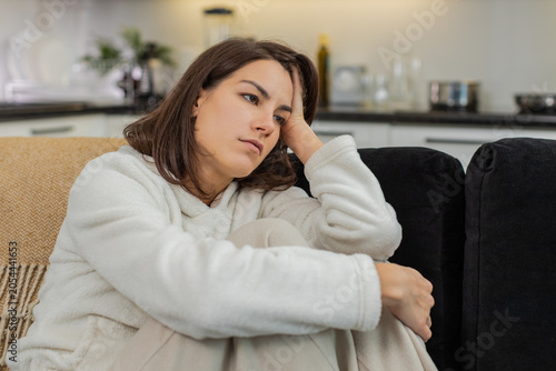 Young woman at home sees negative news on phone feels stress worry and low spirit holds breath. Caucasian girl on sofa hugs knees bows head and waits for support and relief from heavy thought cloud