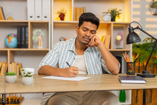 Indian man on home table writes a simple shopping list on paper and plans daily needs with calm focus. Arabian guy on home table makes notes with pen and prepares for local home store trip after work.