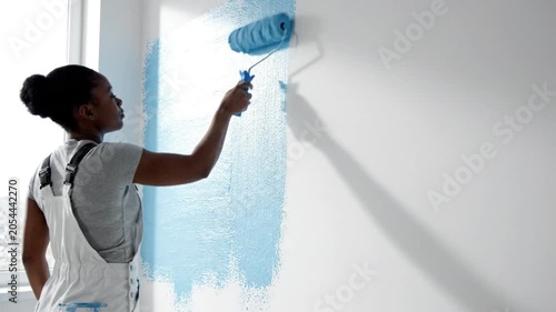 A woman painting a wall blue with a roller in a bright room with a window