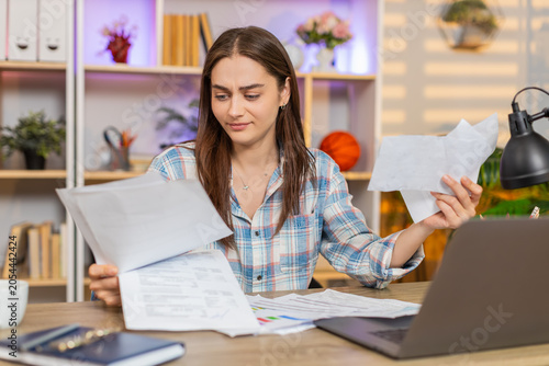 Young woman on home table throws papers after checking bills and receipts feeling angry from high costs. Adult girl on home table shows frustration near laptop during budget work and finance trouble.