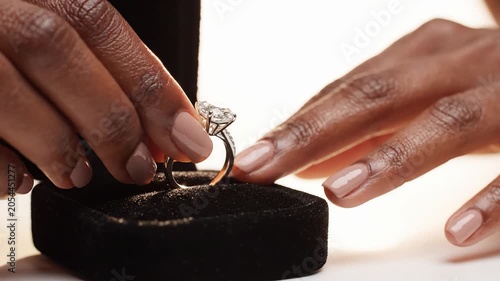 A woman's hand carefully takes a diamond ring out of a black jewelry box on a white surface in this close-up video.