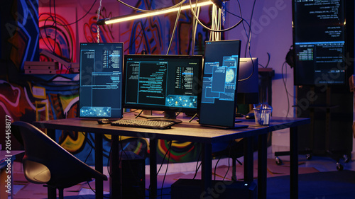 Foto Jib down shot of advanced technology computer system running malicious code in empty warehouse, PC monitors in empty graffiti painted underground hideout used by hackers to commit illicit activities