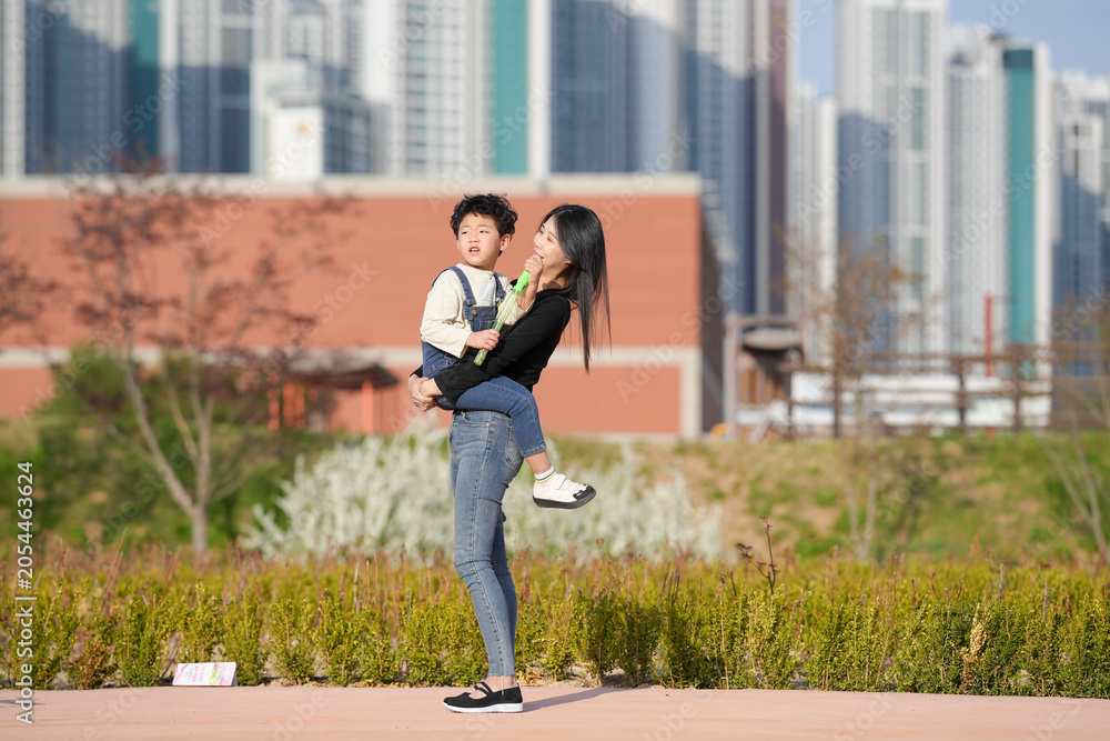 Fototapeta premium Mother hugs child warmly in urban park. Sunlight glows on their joyful faces. Buildings rise softly behind greenery. Perfect moment of love, play, and peace. Great for family, lifestyle