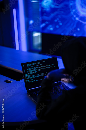Mysterious hacker in black hoodie stealing confidential data on laptop. Cyber criminal coding dangerous malware virus to attack network system in dark blue room.
