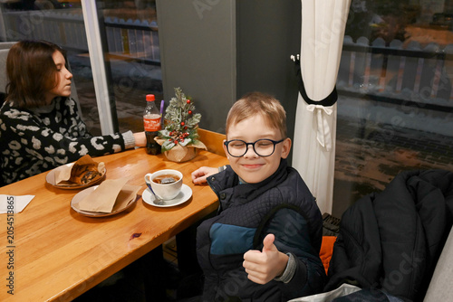 Fotografija boy wearing glasses showing thumb up while sitting at a table in a cafe