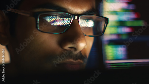 Close-up of a man wearing glasses reflecting computer code in a dark room.