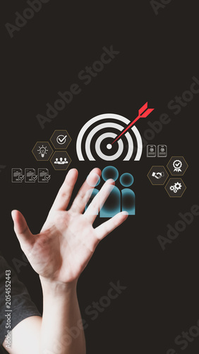 A person's hand reaches toward a virtual interface displaying a target bullseye with an arrow, user icons, and hexagonal business graphics on a dark background.