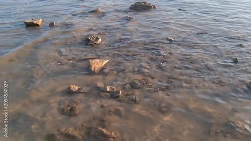 Small, clear sea waves gently washing over scattered brown rocks on a shallow sandy shore during daytime.