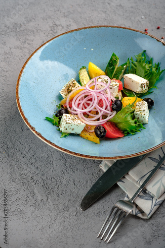 Fresh Greek salad with feta cheese cubes, black olives, cherry tomatoes, cucumber, bell pepper, red onion rings and lettuce in light blue bowl. Classic Mediterranean horiatiki salad