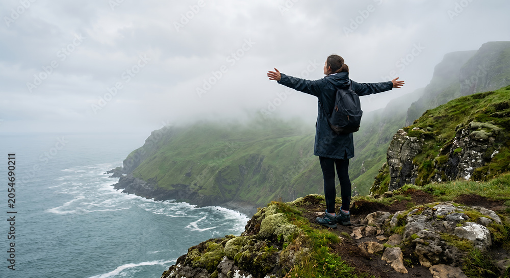 Obraz premium A hiker stands on a cliff edge with arms outstretched, overlooking the ocean and misty green mountains under an overcast sky.
