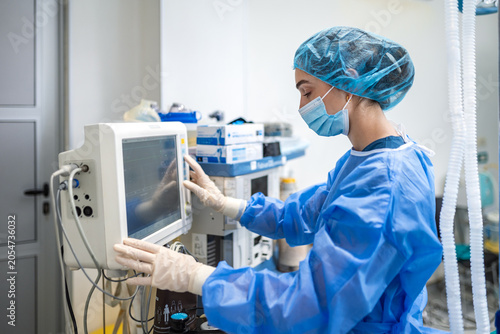 Female anesthesiologist checking medical monitor screen in operating room during surgery. Professional doctor in blue protective uniform, mask and gloves adjusting anesthesia ventilation machine.