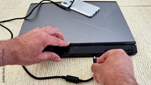 Hands connect cable to back of metallic gray laptop with angular design, smartphone resting on top, showcasing modern tech setup for charging or data transfer on textured table