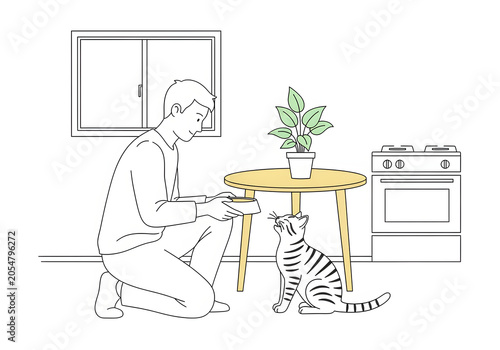 Young man kneeling on a kitchen floor feeding his cute striped pet cat a bowl of fresh food near a small wooden table