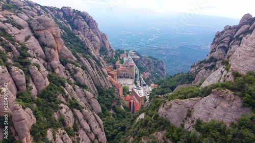 Overhead view down the valley of Montserrat monastery complex flanked by dramatic vertical conglomerate rock formations, with the misty Catalan plain stretching into the distance