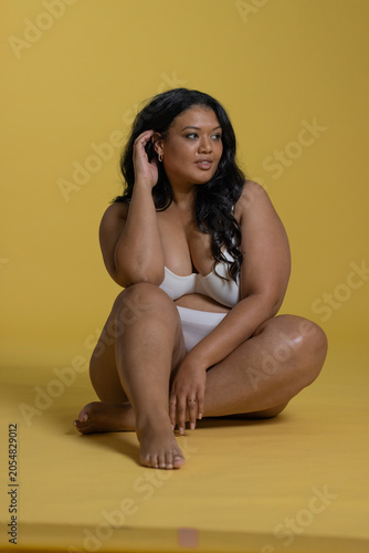 Mid-adult woman sitting cross-legged in studio on yellow backdrop in white underwear touching hair