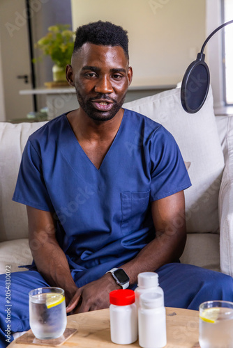 Male medical worker wearing navy scrubs speaking into microphone at home studio with meds bottles