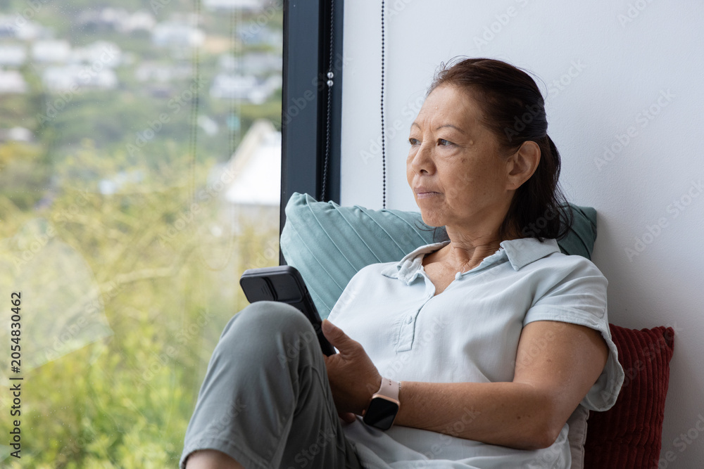 Fototapeta premium Senior Asian woman sitting on window seat, holding smartphone, showing smartwatch, copy space