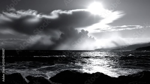 Dramatic monochrome seascape showing a stormy ocean with sunlight shining through a dense cloud formation over rocky shoreline.