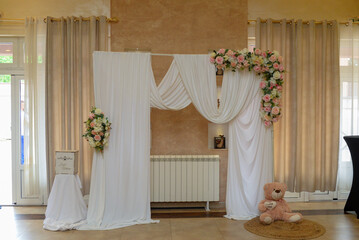 Decoration as a background for photos with the newlyweds. A white curtain, bouquets of roses, a teddy bear holding a heart with the words "I love you" and a gift box with best wishes written on it