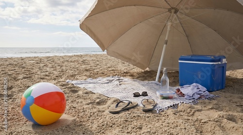 Beige beach umbrella protecting a striped towel with a blue cooler and colorful beach ball