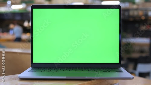Laptop with green screen sits on wooden table. Background shows blurred cafe with people moving. Focus stays on laptop throughout. Ideal for presentations, tutorials, or video editing