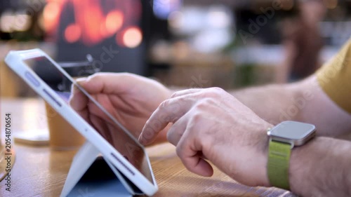 Close-up of hands using tablet on wooden table. Person wears smartwatch with green strap. Background softly blurred, cafe or workspace ambiance. Fingers tap and swipe screen actively