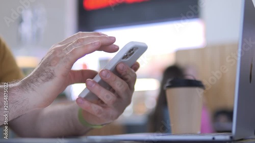 Person uses smartphone while sitting at table. Laptop and coffee cup sit nearby. Background shows cafe with blurred people. Hands scroll phone screen actively
