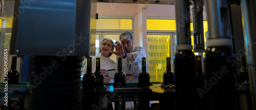Food and Beverage Industry Workers in White Lab Coats and Hairnets Using Digital Tablets to Monitor and Inspect Automated Wine Bottling Production Line in Modern Winery Manufacturing Facility