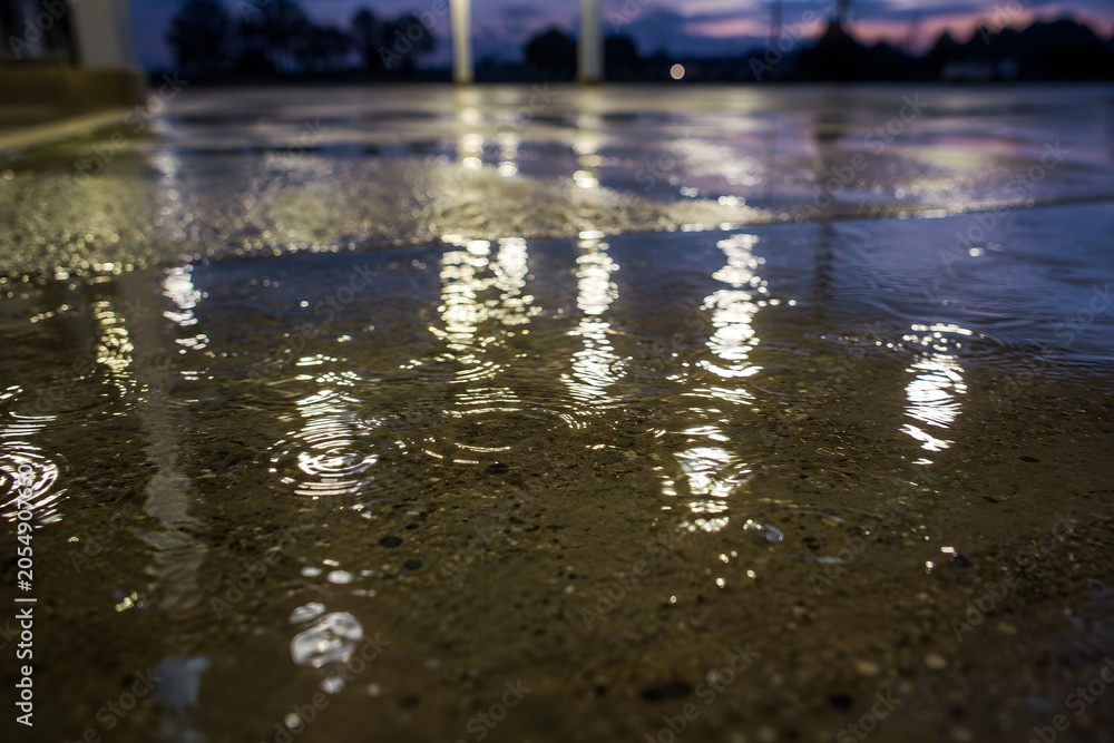 Fototapeta premium Rain Puddles On Concrete Gas Station Forecourt Reflecting Canopy Lights At Dusk With Ripples Detail
