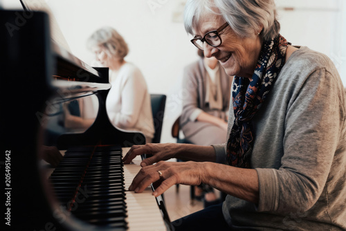 Elderly woman plays grand piano with joy. Senior lady enjoys classical music with friends nearby. Her hands move across keys making beautiful sound.